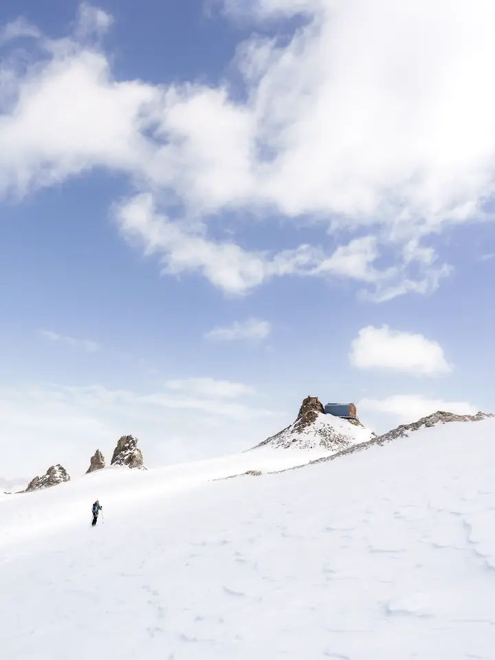 Alpiniste solitaire progressant dans une neige immaculée vers un refuge perché, sous un ciel de haute montagne — série photographique Face à Face